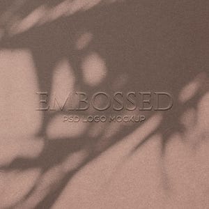 Pode incluir: Close-up de uma superfície castanha texturizada com a palavra "EMBOSSED" em relevo no centro. Abaixo, o texto "PSD LOGO MOCKUP". As sombras da folhagem são projetadas sobre a superfície.