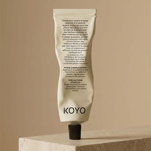 May include: A tube of KOYO sunscreen with a black cap, set against a neutral background. The tube is off-white with text in French. The sunscreen is designed for sensitive skin and offers protection against sun damage.