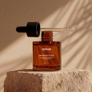 Puede incluir: Una botella de suero de vidrio marr&oacute;n con un cuentagotas negro, etiquetada como "SERUM" e "INFINIUM GLOW". La botella se encuentra sobre una superficie de piedra texturizada. El cuentagotas est&aacute; parcialmente insertado en la botella, con una pipeta de vidrio transparente.