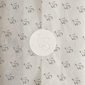 May include: White tissue paper with a repeating pattern of a stylized "IS" logo in gray. A white circular sticker with an embossed "IS" logo is centered on the paper. The paper is folded, creating vertical lines.