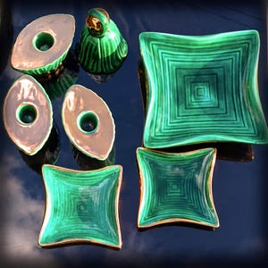 May include: An assortment of decorative ceramic pieces in shades of green and gold. The collection includes oval-shaped candle holders, square dishes with concentric square patterns, and a small, rounded figurine. The pieces have a glossy finish.