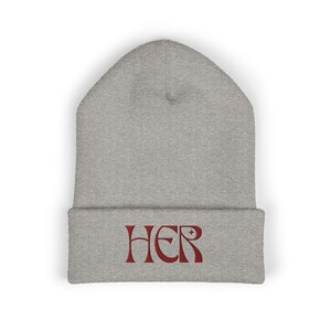May include: A grey beanie hat with the word "HER" embroidered in reddish-brown. The hat is made of a soft, knitted material and has a folded brim. The text is centered on the brim.