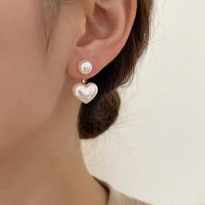 May include: A pair of pearl earrings. Each earring has a small, round pearl stud at the top and a larger, heart-shaped pearl pendant below. The earrings are shown against a person's ear and shoulder.