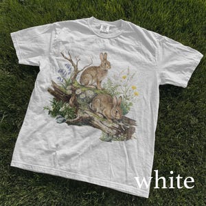 K&ouml;nnte beinhalten: Wei&szlig;es T-Shirt mit einer detaillierten Illustration von zwei braunen Kaninchen auf einem Baumstamm, umgeben von Wildblumen und Gr&uuml;n. Das T-Shirt hat einen klassischen Rundhalsausschnitt und kurze &Auml;rmel. Das Wort "white" ist unten aufgedruckt.