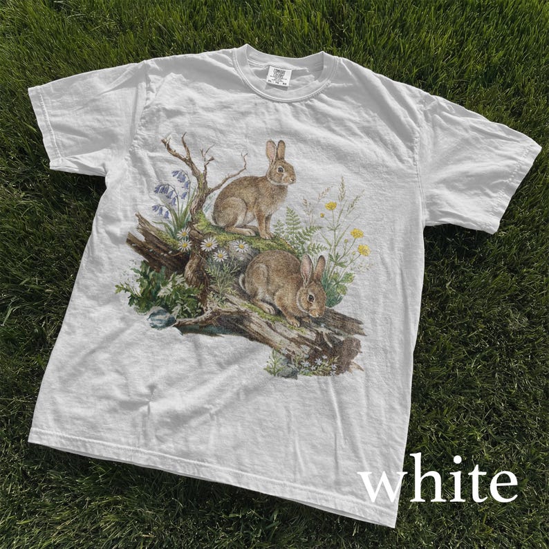 K&ouml;nnte beinhalten: Wei&szlig;es T-Shirt mit einer detaillierten Illustration von zwei braunen Kaninchen auf einem Baumstamm, umgeben von Wildblumen und Gr&uuml;n. Das T-Shirt hat einen klassischen Rundhalsausschnitt und kurze &Auml;rmel. Das Wort "white" ist unten aufgedruckt.
