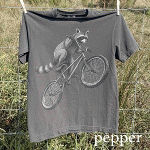Puede incluir: Camiseta gris con un mapache montando en bicicleta, con la palabra "pepper" impresa en la parte inferior. El mapache está representado en escala de grises. La camiseta está colgada en un tendedero, con un fondo natural.