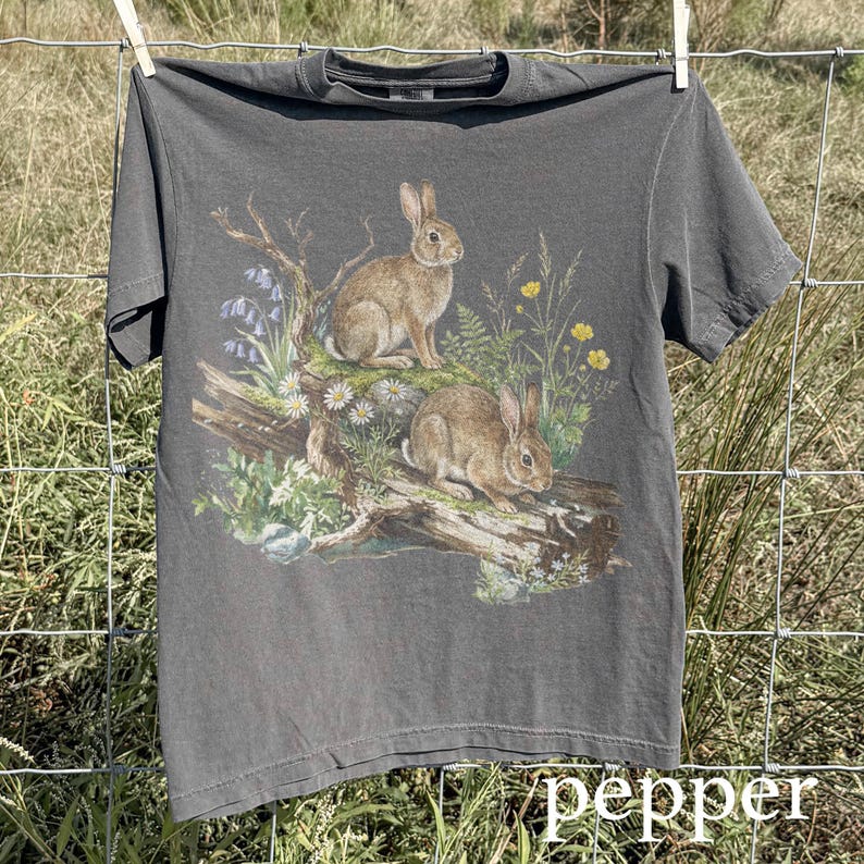 K&ouml;nnte beinhalten: Graues T-Shirt mit einer detaillierten Illustration von zwei Kaninchen auf einem Baumstamm, umgeben von Wildblumen und Bl&auml;ttern. Das Wort "pepper" ist in wei&szlig;er Schrift unten aufgedruckt. Das T-Shirt h&auml;ngt an einer W&auml;scheleine.