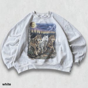 May include: White sweatshirt featuring a graphic of three wolves in a forest scene under a moonlit sky with lightning. The wolves are depicted in shades of brown, white, and gray. The word "white" is printed at the bottom of the sweatshirt.