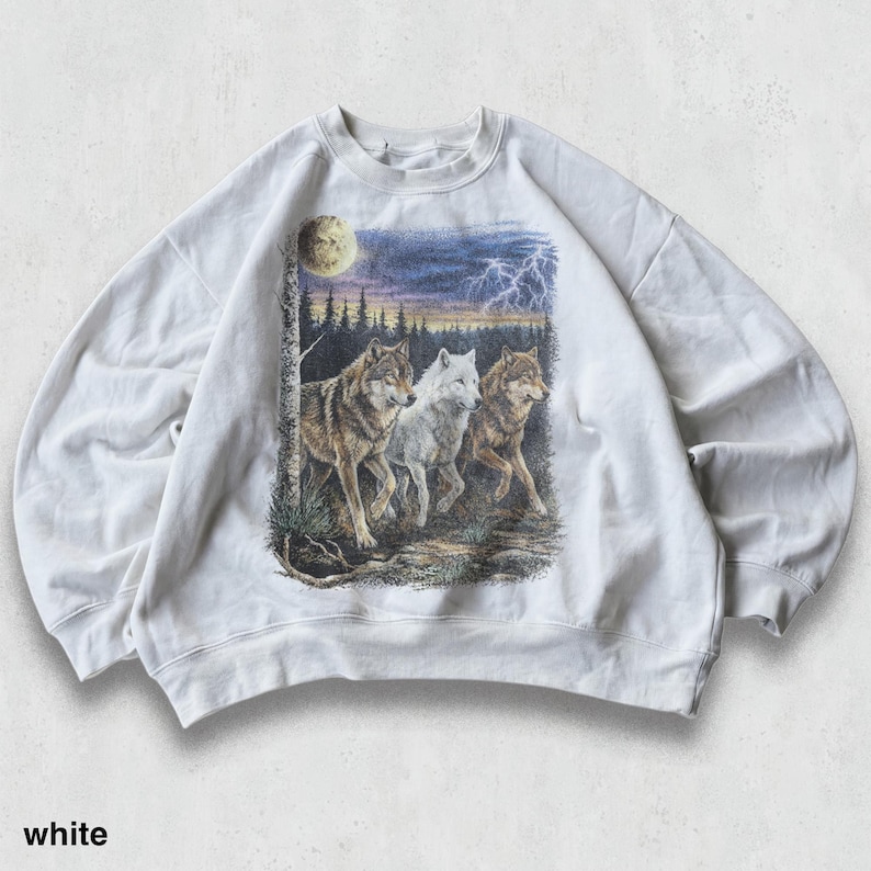 May include: White sweatshirt featuring a graphic of three wolves in a forest scene under a moonlit sky with lightning. The wolves are depicted in shades of brown, white, and gray. The word "white" is printed at the bottom of the sweatshirt.