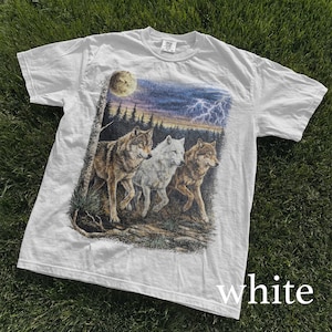 May include: White t-shirt featuring a graphic of three wolves in a forest setting under a moonlit sky with lightning. The wolves are depicted in shades of brown and white. The word "white" is printed at the bottom of the shirt.