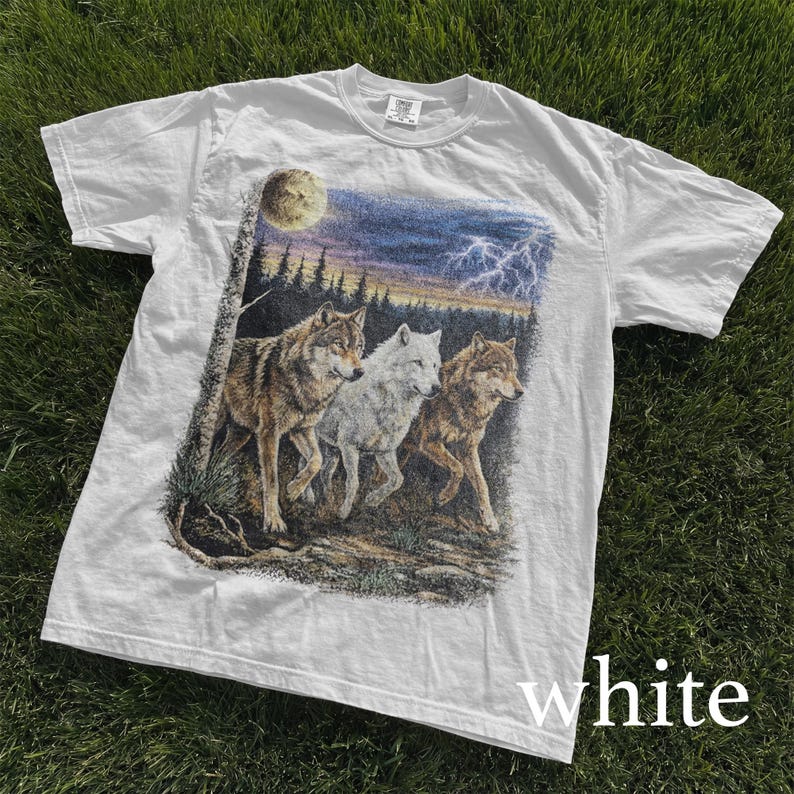 May include: White t-shirt featuring a graphic of three wolves in a forest setting under a moonlit sky with lightning. The wolves are depicted in shades of brown and white. The word "white" is printed at the bottom of the shirt.