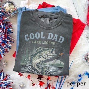 May include: A stack of folded t-shirts in red, white, and blue. The top shirt is dark gray with the text "COOL DAD LAKE LEGEND" and a graphic of a fish jumping out of the water. The shirts are on a white, fluffy surface.
