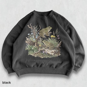 May include: Black crewneck sweatshirt featuring a detailed illustration of two frogs on a log with colorful wildflowers and foliage. The word "black" is printed at the bottom. The design is printed on a dark gray, soft fabric.