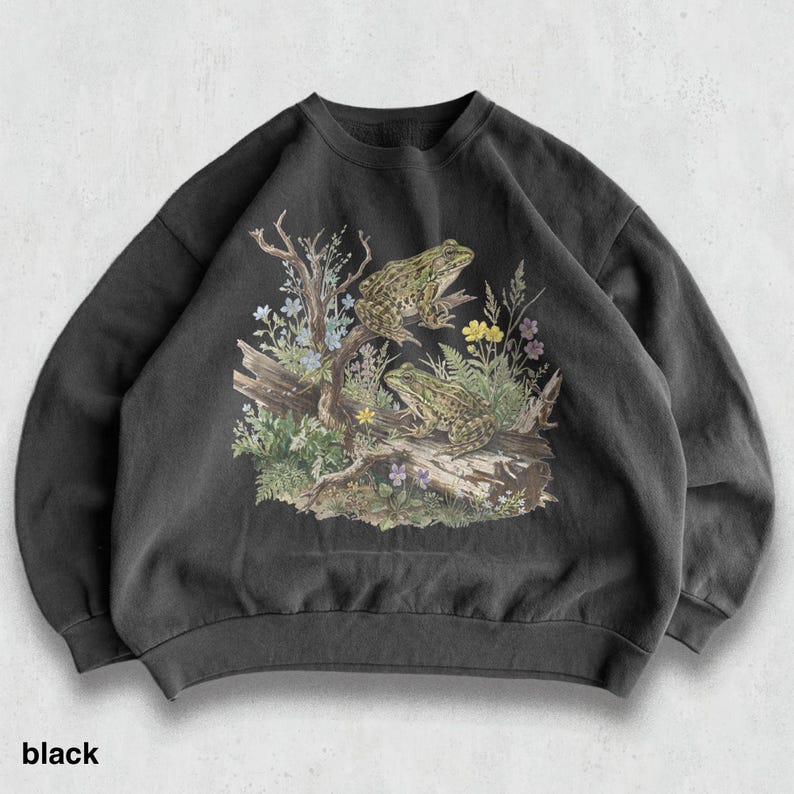 May include: Black crewneck sweatshirt featuring a detailed illustration of two frogs on a log with colorful wildflowers and foliage. The word "black" is printed at the bottom. The design is printed on a dark gray, soft fabric.