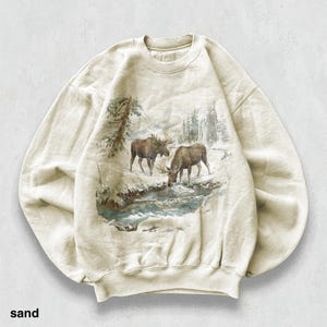 May include: Sand-coloured sweatshirt with a detailed graphic of two moose in a snowy landscape, drinking from a stream. The design includes trees and a winter scene. The word "sand" is printed at the bottom.