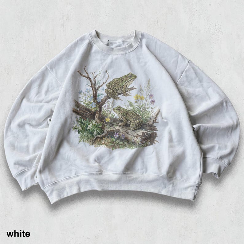 May include: White sweatshirt featuring a detailed illustration of two frogs perched on a log with foliage. The graphic includes a tree branch, flowers, and ferns. The word "white" is printed at the bottom.