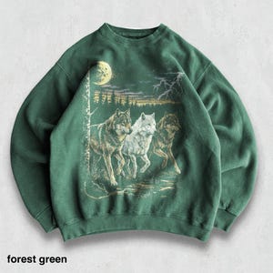 May include: Forest green sweatshirt featuring a graphic of three wolves running through a forest under a full moon. The design includes a detailed illustration of trees and a lightning bolt. The text "forest green" is at the bottom.