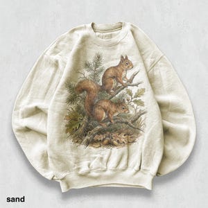 Puede incluir: Sudadera color arena con una ilustración detallada de dos ardillas posadas en una rama de árbol con hojas y bellotas. El gráfico está en tonos marrones y verdes, sobre un fondo neutro. La palabra "sand" está impresa en la parte inferior.