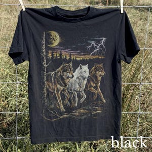 May include: Black t-shirt featuring a graphic of three wolves running under a moonlit sky with lightning. The wolves are depicted in shades of brown and white. The word "black" is printed at the bottom of the shirt.