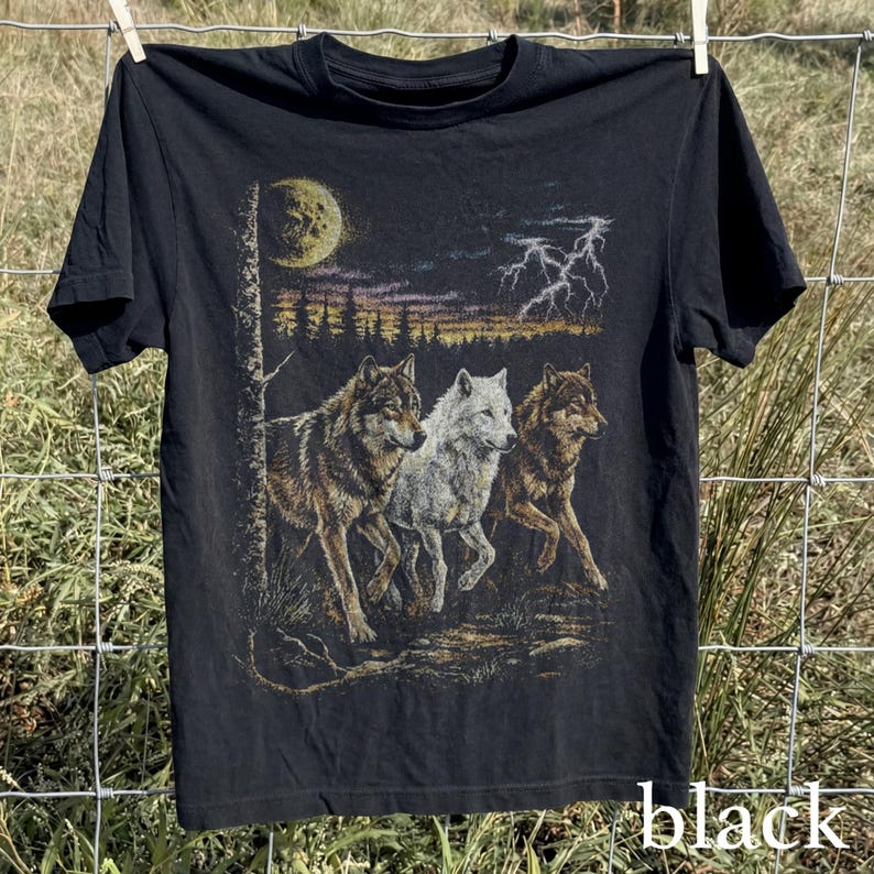 May include: Black t-shirt featuring a graphic of three wolves running under a moonlit sky with lightning. The wolves are depicted in shades of brown and white. The word "black" is printed at the bottom of the shirt.