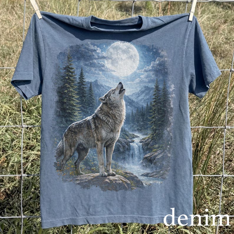 May include: Denim blue t-shirt featuring a detailed graphic of a howling wolf under a full moon. The design includes a waterfall, mountains, and evergreen trees. The word "denim" is printed at the bottom.