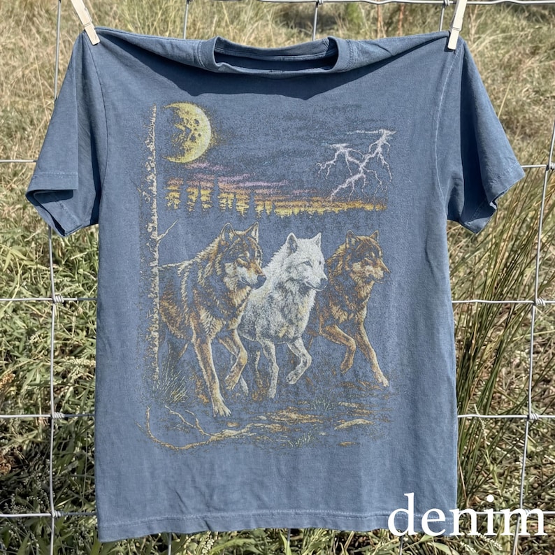 May include: Blue denim t-shirt featuring a graphic of three wolves in a nighttime landscape with a moon and lightning. The shirt is hanging on a clothesline. The word "denim" is printed at the bottom.