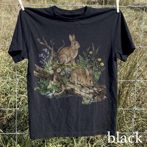 K&ouml;nnte beinhalten: Schwarzes T-Shirt mit einer detaillierten Illustration von zwei braunen Kaninchen auf einem Baumstamm, umgeben von Wildblumen und Bl&auml;ttern. Das Wort "black" ist unten auf dem T-Shirt aufgedruckt.