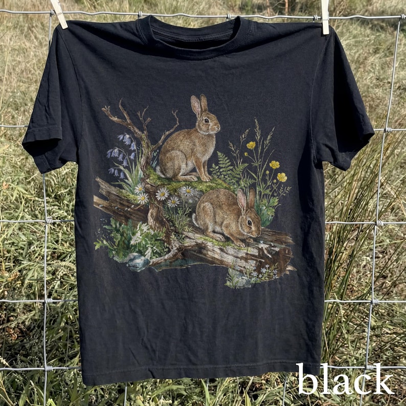 K&ouml;nnte beinhalten: Schwarzes T-Shirt mit einer detaillierten Illustration von zwei braunen Kaninchen auf einem Baumstamm, umgeben von Wildblumen und Bl&auml;ttern. Das Wort "black" ist unten auf dem T-Shirt aufgedruckt.