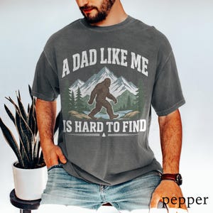 Pode incluir: Camiseta cinza escuro com o texto "A DAD LIKE ME IS HARD TO FIND" em branco. O gráfico apresenta um Bigfoot caminhando em frente a montanhas e árvores. A camisa é um estilo confortável e casual.