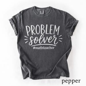 Könnte beinhalten: Ein anthrazitfarbenes Kurzarm-T-Shirt mit Vintage-Waschung. Auf der Vorderseite befindet sich der weiße Schriftzug "PROBLEM solver" in einer Mischung aus Block- und Schreibschrift, darunter der Hashtag "#mathteacher" in einer kleineren serifenlosen Schrift.