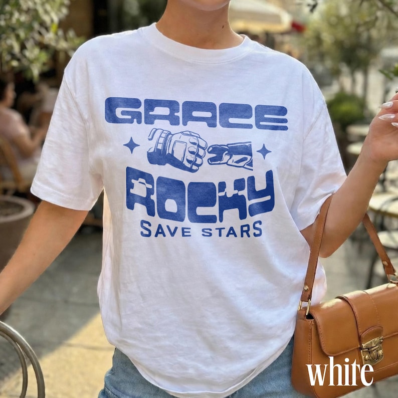 May include: White t-shirt with a blue graphic design. The design includes the words "GRACE ROCKY SAVE STARS" in a bold, stylized font, along with a graphic of a fist. The shirt is short-sleeved and appears to be made of cotton.