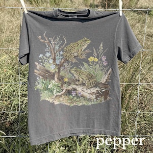 May include: A gray t-shirt featuring a detailed illustration of two frogs perched on a log with wildflowers and foliage. The word "pepper" is printed in white at the bottom right corner. The shirt is hanging on a clothesline.
