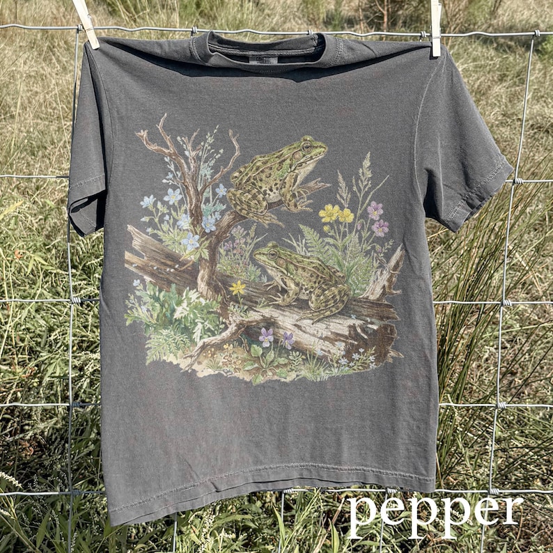 May include: A gray t-shirt featuring a detailed illustration of two frogs perched on a log with wildflowers and foliage. The word "pepper" is printed in white at the bottom right corner. The shirt is hanging on a clothesline.