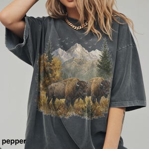 May include: Dark grey t-shirt featuring a scenic landscape with two bison in a field, mountains, trees, and birds. The word "pepper" is printed at the bottom left.