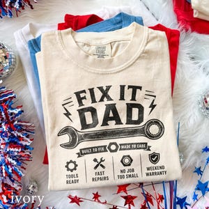 May include: Ivory-colored t-shirt with the text "FIX IT DAD" and a wrench graphic. Additional text includes "BUILT TO FIX," "MADE TO CARE," and icons for tools, repairs, and weekend warranty. Other shirts in white, blue, and red are visible.