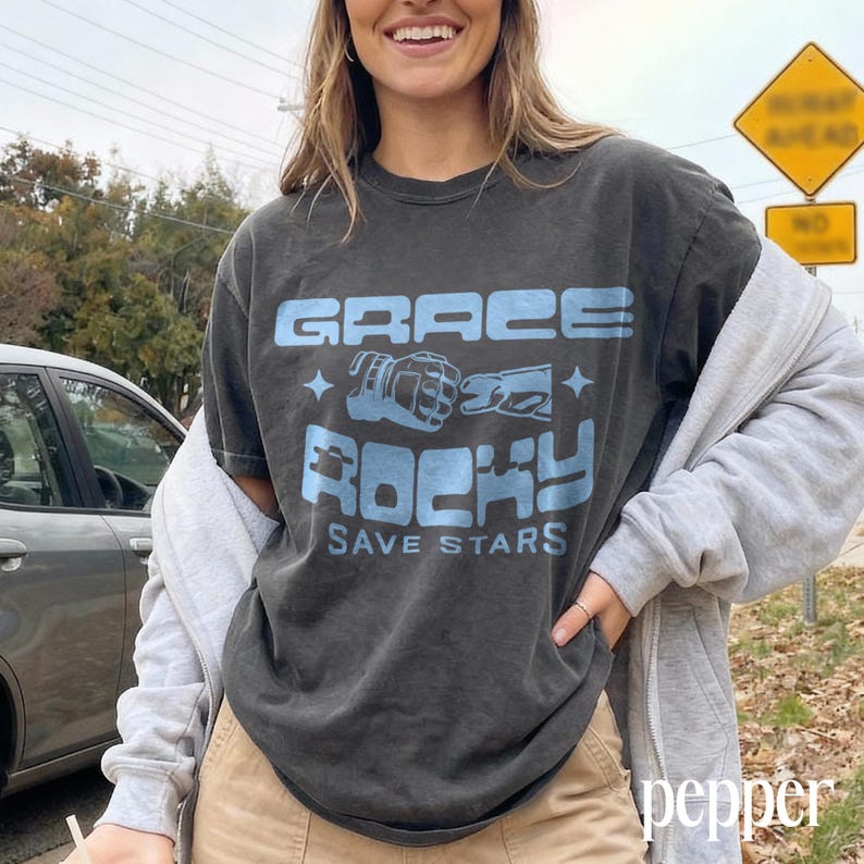 May include: Dark gray t-shirt with the text "GRACE ROCKY SAVE STARS" in light blue. The shirt features a graphic of boxing gloves and stars. The model is wearing a gray zip-up hoodie and tan pants.