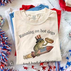Könnte beinhalten: Elfenbeinfarbenes T-Shirt mit dem Text "Running on hot dogs and bad decisions". Die Grafik zeigt einen Frosch mit patriotischem Hut, der einen Hotdog hält und auf einer Rakete reitet. Weitere rote, weiße und blaue T-Shirts sind dahinter gestapelt.
