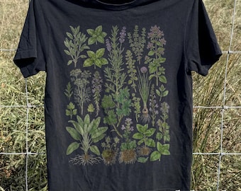 May include: Black t-shirt featuring a botanical illustration of various herbs and plants, including basil, lavender, and rosemary. The design showcases detailed drawings of the plants with their roots, stems, and leaves. The word "black" is printed at the bottom right.