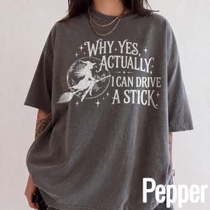 Könnte beinhalten: Ein anthrazitfarbenes Kurzarm-T-Shirt mit einer weißen Grafik einer Hexe, die auf einem Besenstiel vor einem Mond fliegt. Der Text lautet: WHY YES, ACTUALLY I CAN DRIVE A STICK. Das T-Shirt hat einen Rundhalsausschnitt und eine lockere Passform.