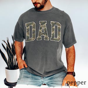 以下が含まれることがあります： 胸に迷彩柄で「DAD」の文字が入ったグレーのTシャツ。Tシャツは半袖でクルーネックです。モデルはブルージーンズと茶色の腕時計を着用しています。