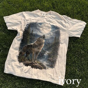 May include: Ivory-colored t-shirt featuring a detailed graphic of a wolf howling at the moon in a forest setting. The design includes a waterfall and trees. The shirt is made of a soft material.
