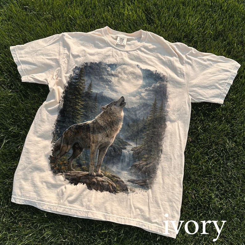 May include: Ivory-colored t-shirt featuring a detailed graphic of a wolf howling at the moon in a forest setting. The design includes a waterfall and trees. The shirt is made of a soft material.