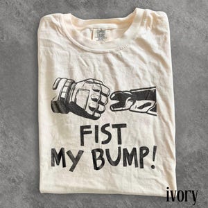 Op de afbeelding: Ivoor kleurig t-shirt met een zwarte afbeelding van twee gehandschoende handen die een vuist geven. De tekst "FIST MY BUMP!" staat eronder gedrukt. Het shirt is gemaakt van een zacht materiaal en heeft een ronde hals.
