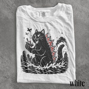May include: A white t-shirt featuring a black graphic illustration of a large cat resembling a monster rising from the ocean waves. The cat holds a small ship, with airplanes flying above, a city skyline in the background, and red spikes.