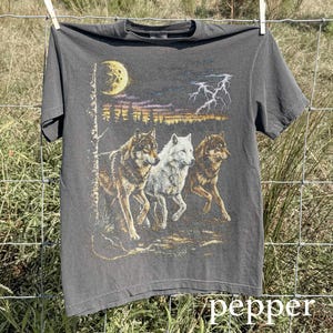 May include: A dark gray t-shirt featuring a graphic of three wolves running under a moonlit sky with lightning. The wolves are in shades of brown and white. The word "pepper" is at the bottom of the shirt.