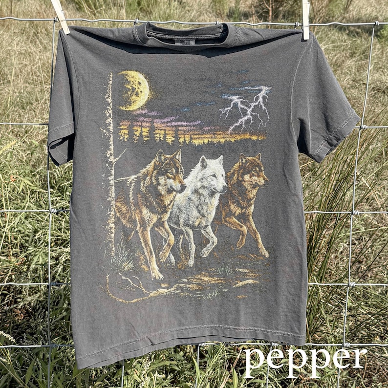 May include: A dark gray t-shirt featuring a graphic of three wolves running under a moonlit sky with lightning. The wolves are in shades of brown and white. The word "pepper" is at the bottom of the shirt.