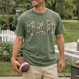 以下が含まれることがあります： 迷彩柄の「DAD」の文字が入ったオリーブグリーンのTシャツ。文字は白い縁取り。茶色と白のアメリカンフットボールを持っている。Tシャツは半袖です。