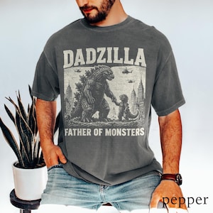 May include: Dark gray t-shirt with a graphic featuring Godzilla and a child Godzilla holding hands, with the text "DADZILLA FATHER OF MONSTERS." The design includes city buildings and helicopters.