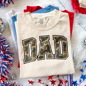 以下が含まれることがあります： 迷彩柄の「DAD」の文字が入ったアイボリー色のTシャツ。赤、青、白の他のTシャツの山の一部です。シャツは、愛国的な装飾が施された白くふわふわした表面にあります。