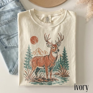 May include: An ivory t-shirt featuring a graphic print of a brown deer standing in a landscape with green pine trees, mountains, and a rust-colored sun. The shirt has a crew neckline and a Comfort Colors brand label printed on the inside collar.